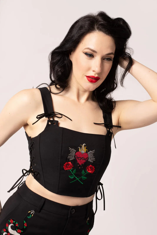 A model wearing a black corset top with embroidered heart and roses on the chest, paired with black shorts adorned with a floral design. The model has a bold red lipstick and is accessorized with black tie details on the corset.
